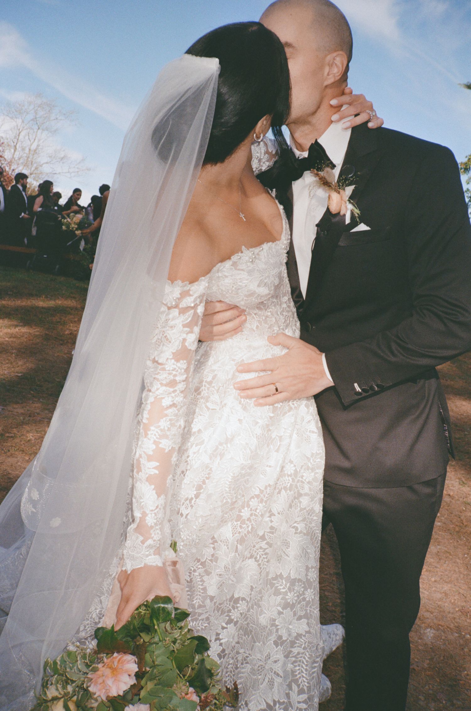A couple shares a romantic kiss at their outdoor wedding ceremony, with the bride in a lace gown and long veil.