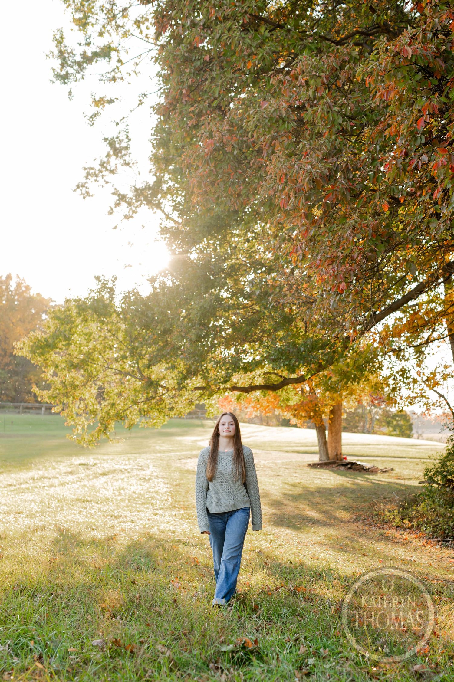 A person in jeans and sweater walks through a sun-lit autumn field under golden trees.