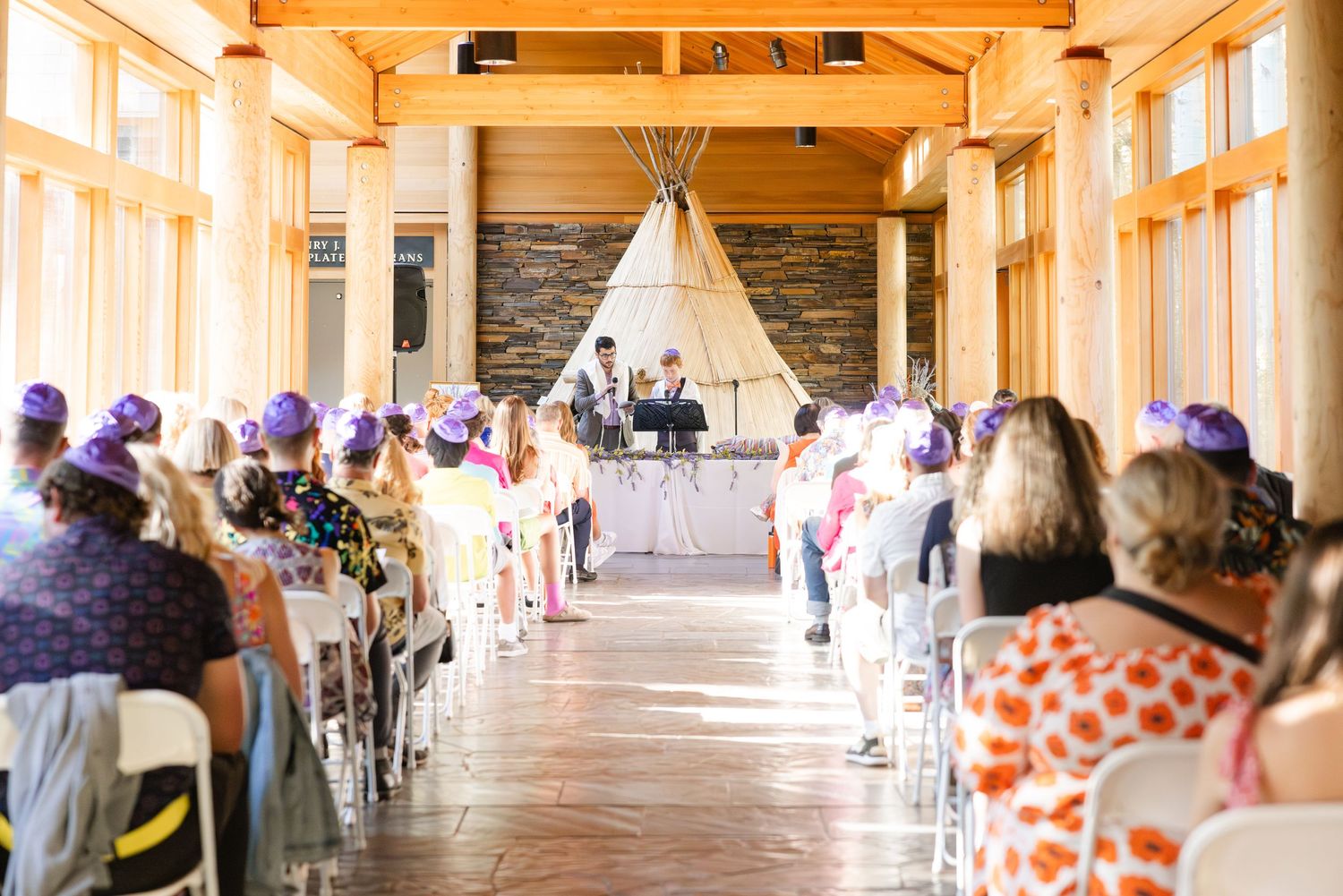 Wedding ceremony setup indoors with a teepee at the altar and guests seated in rustic wooden venue with natural light.