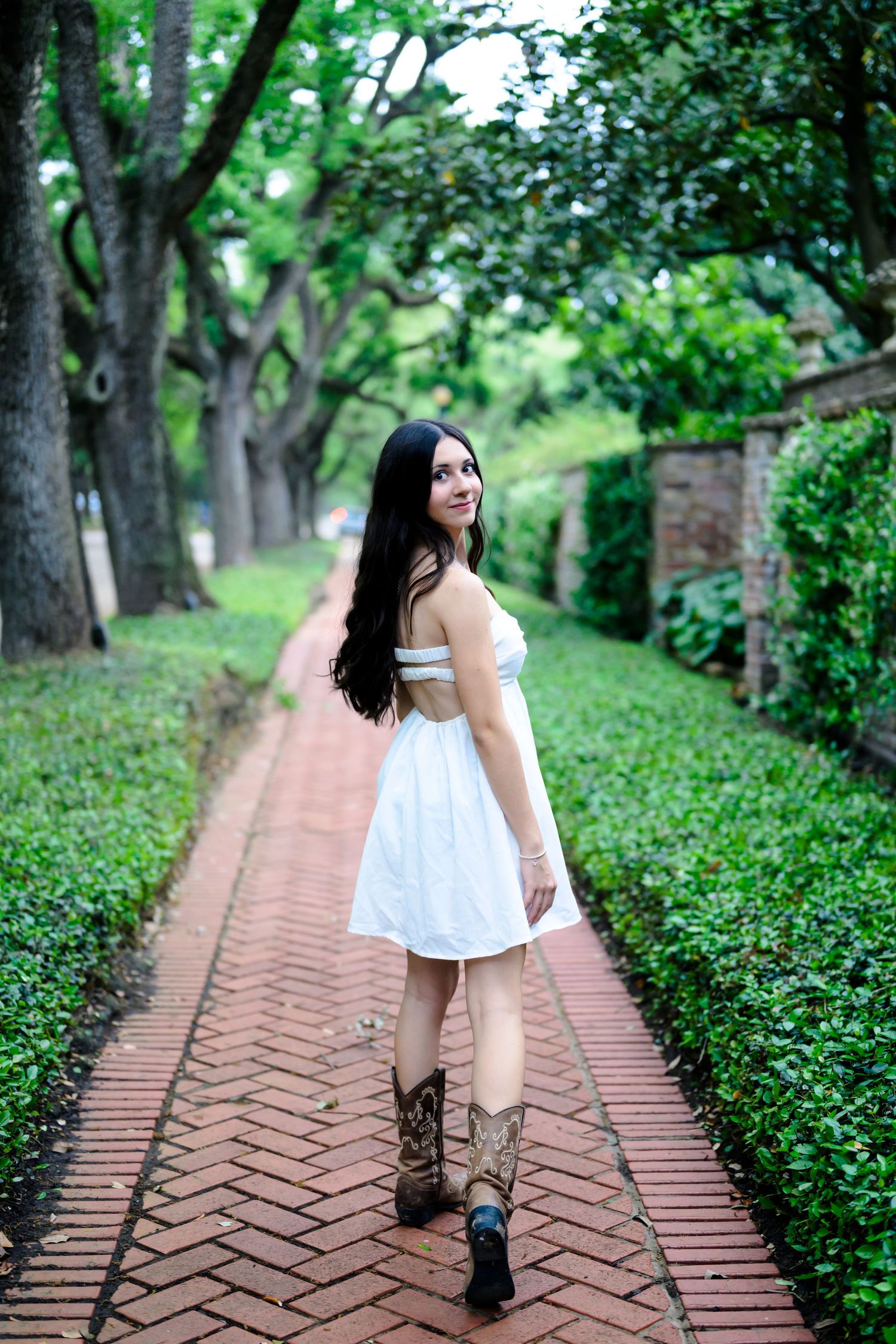 Someone in a white summer dress and brown boots poses on a brick path surrounded by lush green garden scenery.