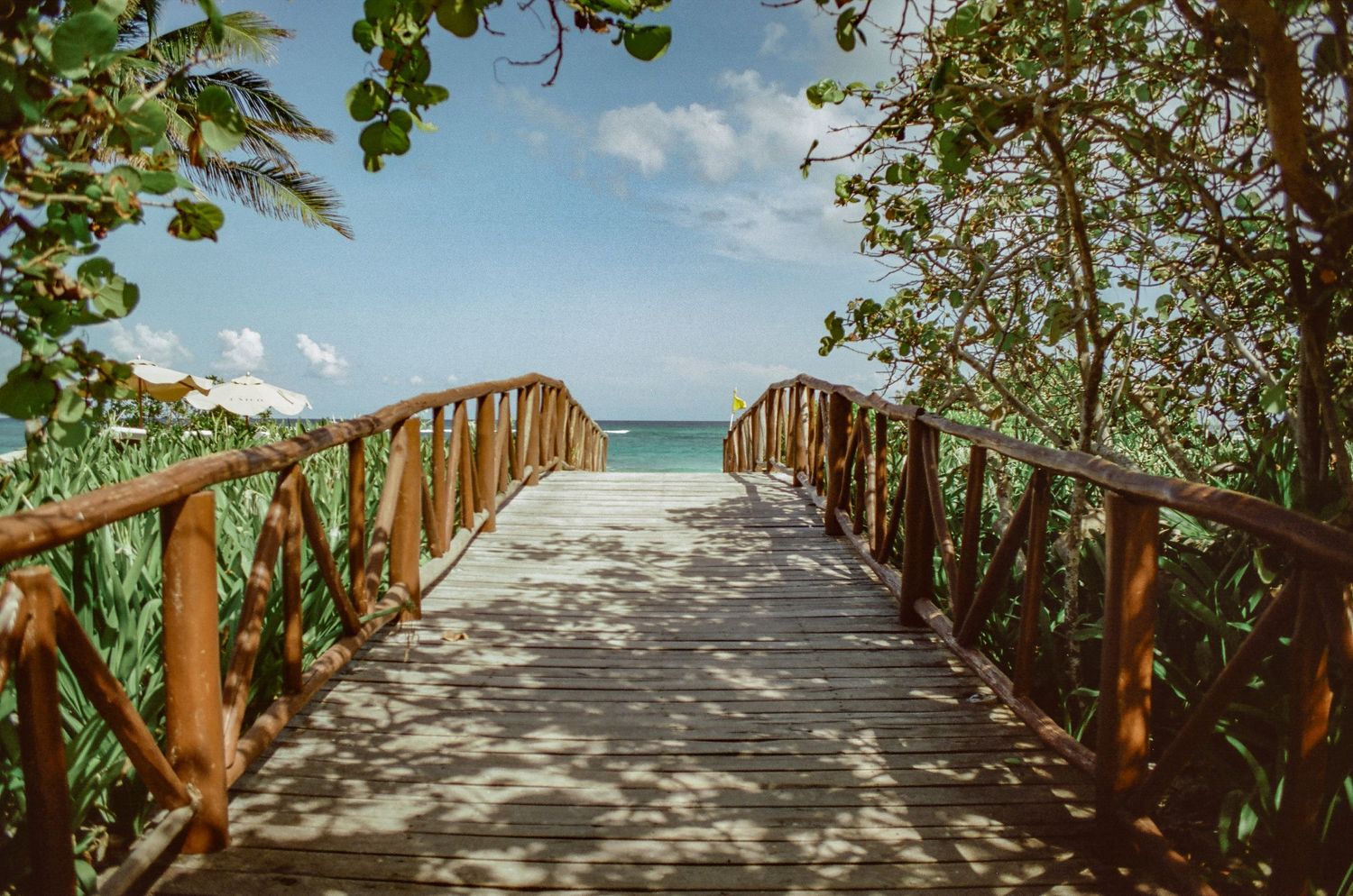 Wooden boardwalk path leading through lush foliage to a pristine tropical beach with turquoise waters and blue sky.
