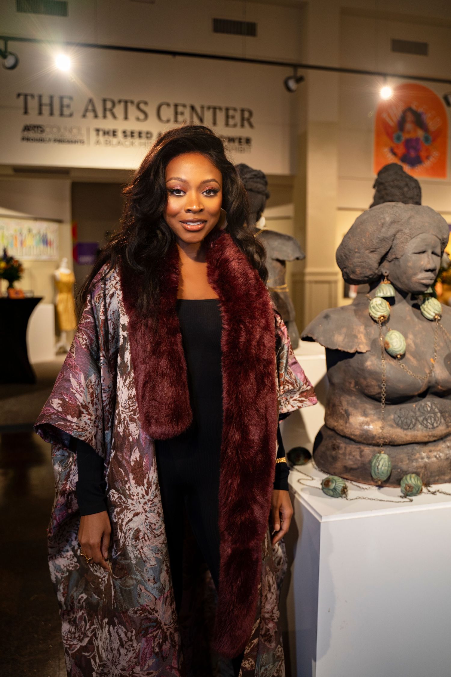 Person wearing burgundy velvet coat standing in arts center gallery next to sculpture display.