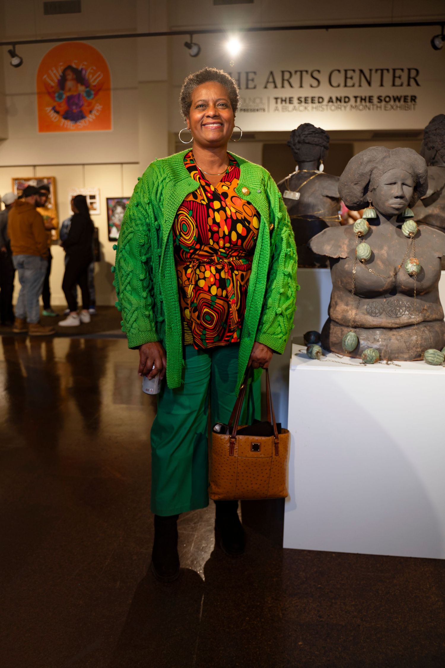 At an arts center exhibition, a figure wears a vibrant green jacket over a patterned orange top with a brown handbag.