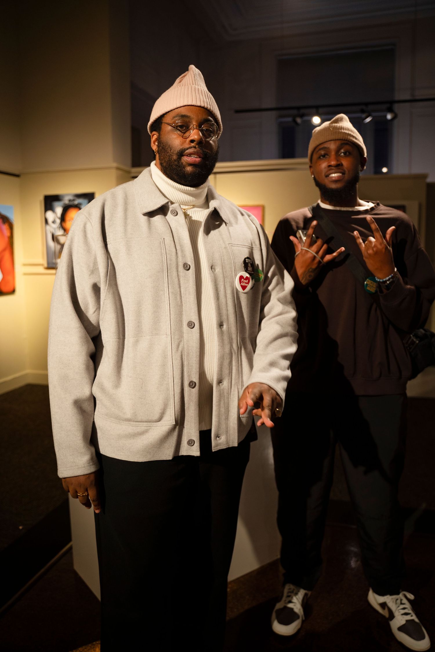 Two people pose together at an art gallery wearing winter attire including beanies and light-colored jackets.