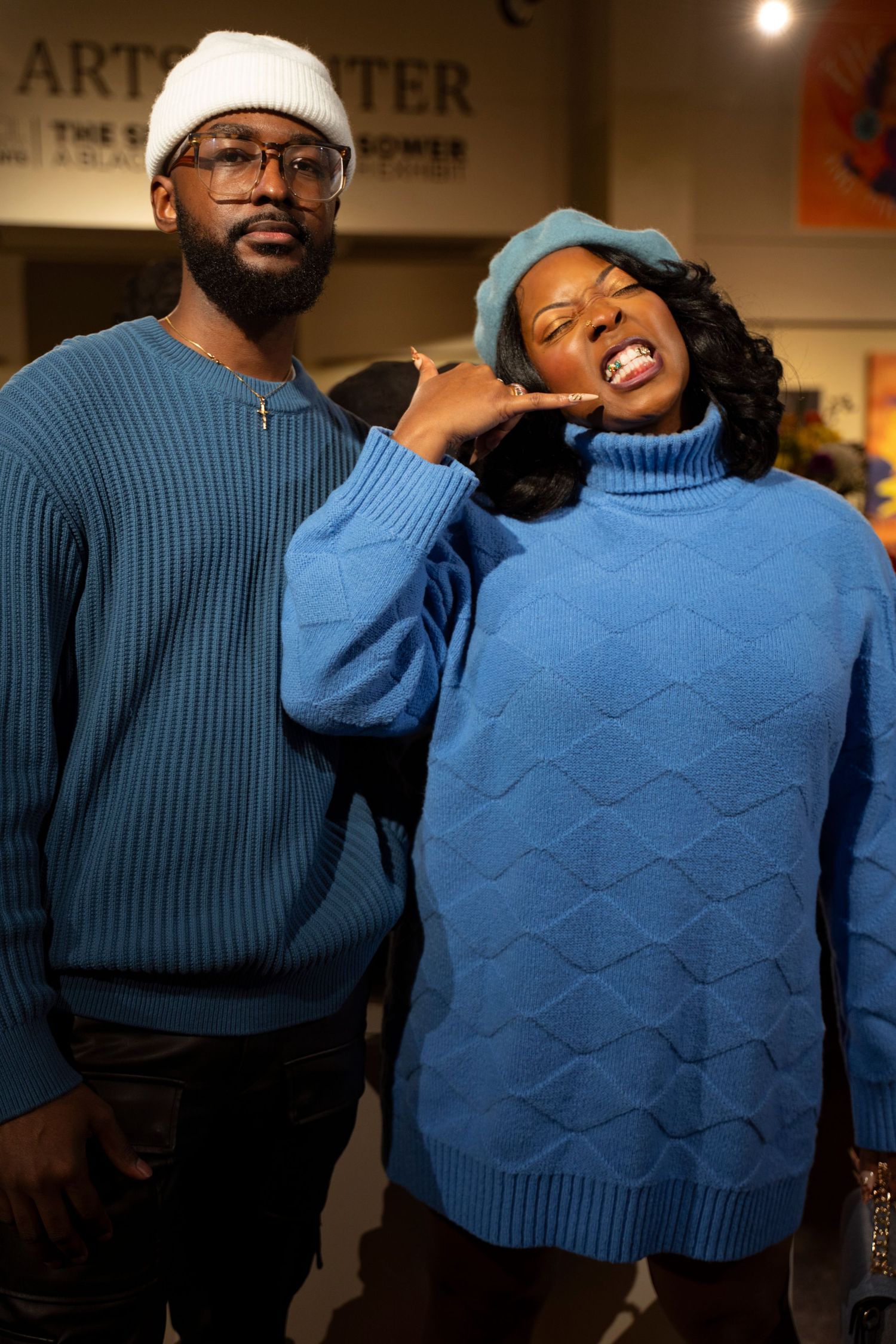 Two people wearing matching blue sweaters pose playfully in an art center gallery setting.