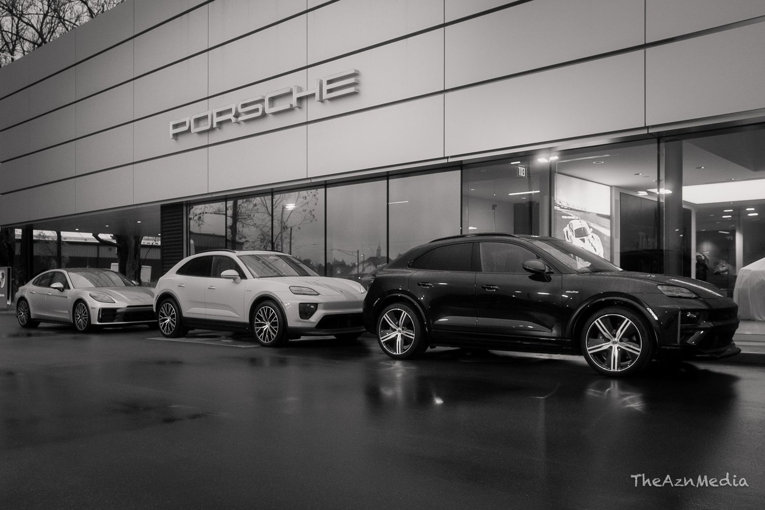 Black and white exterior shot of several Porsche vehicles parked outside a dealership showroom.