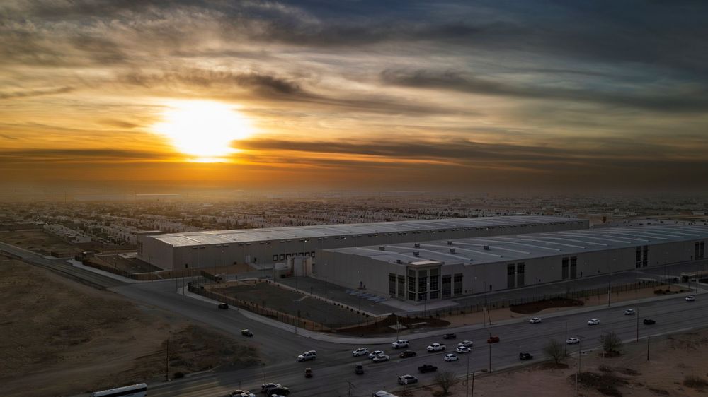 Aerial drone view of a large industrial warehouse complex at sunset with dramatic sky and surrounding roads.