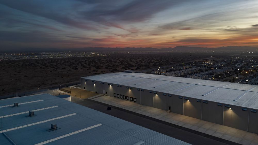 Aerial dusk view of a large industrial warehouse with illuminated exterior lights and city skyline backdrop.
