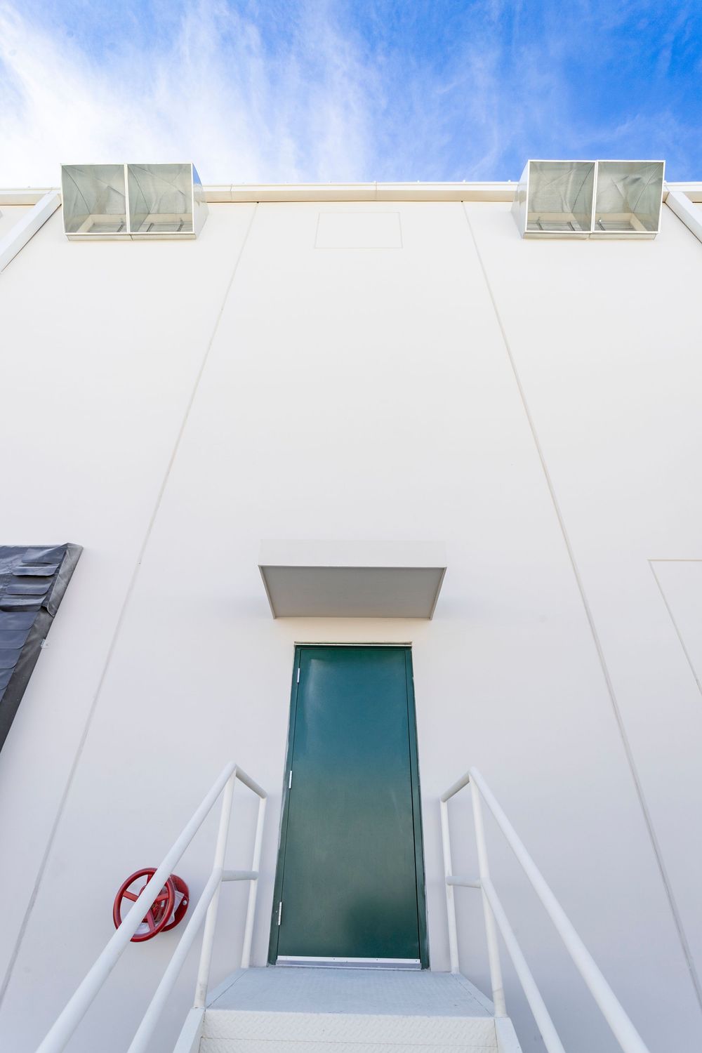 White building exterior with green door, white staircase, red life ring, and blue sky background.