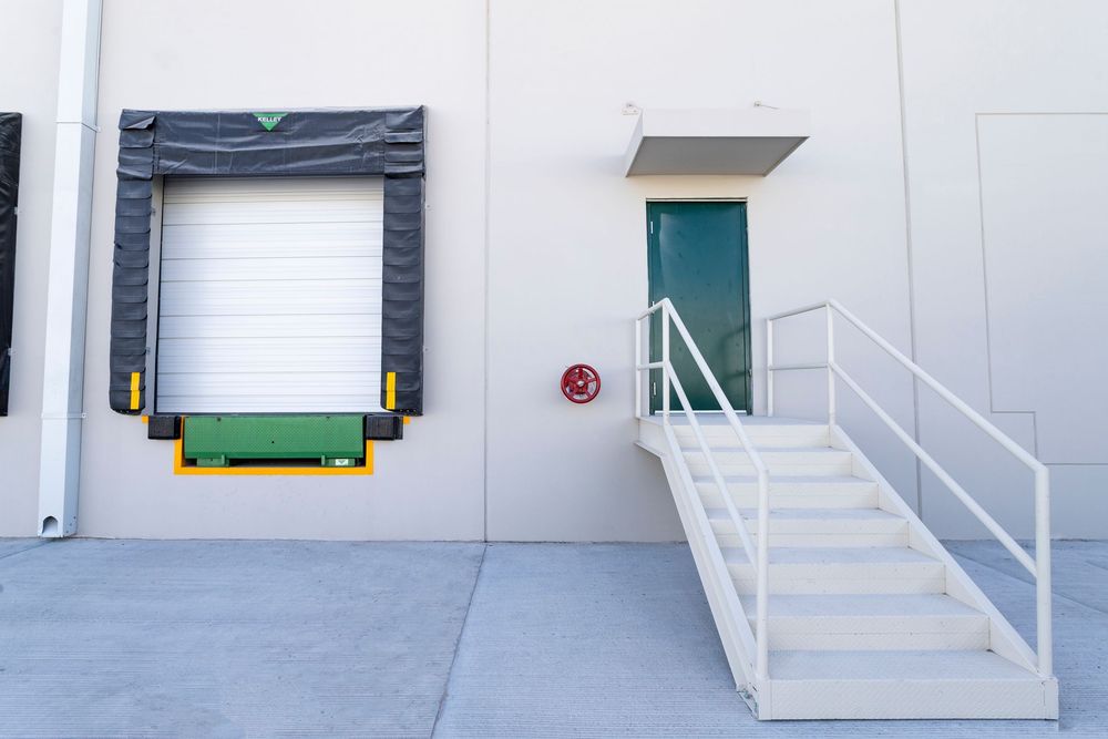 Commercial warehouse exterior with loading dock, roll-up door, green bumpers, and white metal staircase leading to entry door.