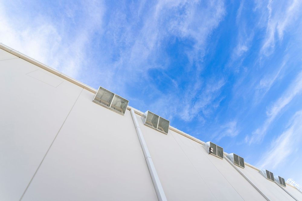 Low-angle view of a modern white building facade against a bright blue sky with wispy clouds.