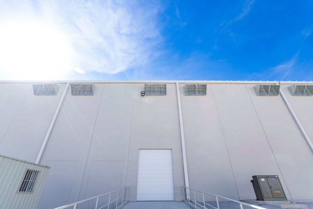 White industrial warehouse exterior with large roll-up door, ventilation units, and bright blue sky backdrop.