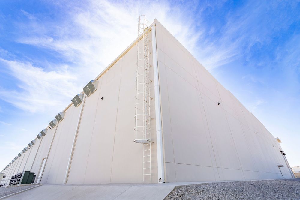 Exterior view of a large modern white industrial warehouse building with a ladder under a bright blue sky.