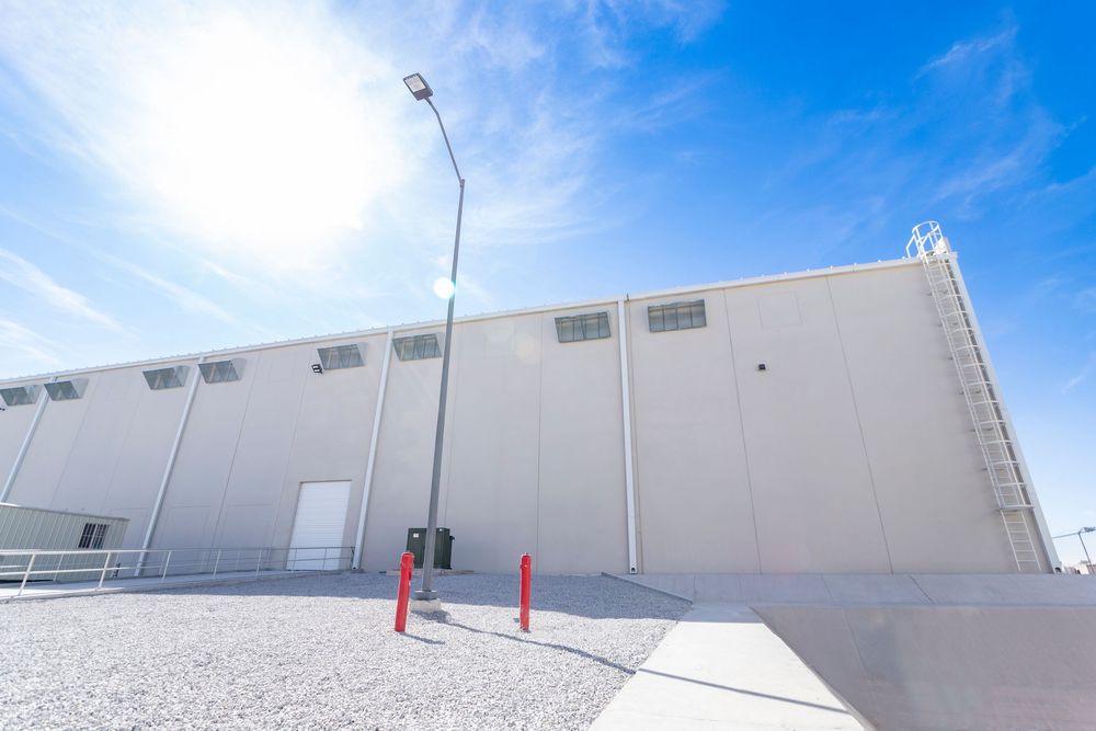Large industrial warehouse exterior with gray metal walls, red bollards, and a street lamp under a bright blue sky.