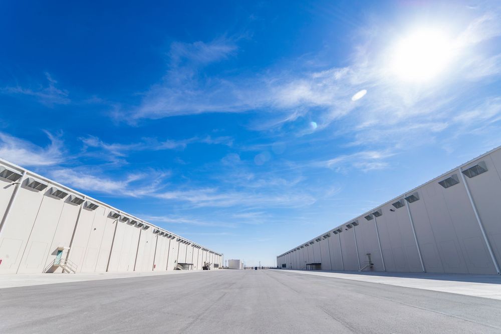Wide-angle view of modern warehouse complex with white walls under bright blue sky with sun flare on clear day.