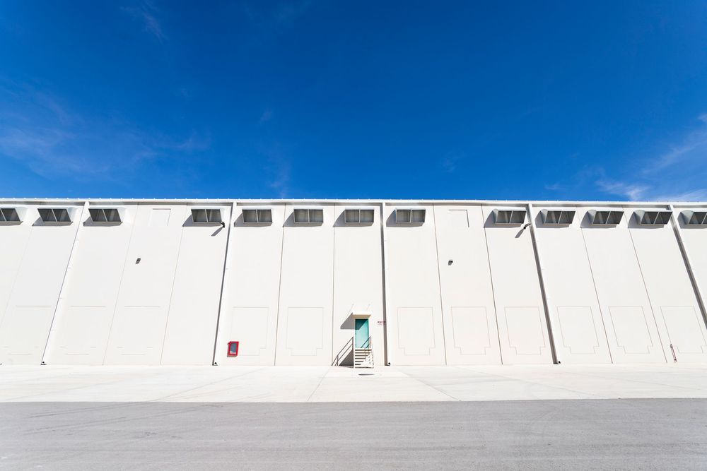 Large white industrial warehouse exterior with blue sky, concrete facade, ventilation units, and a single green door entrance.