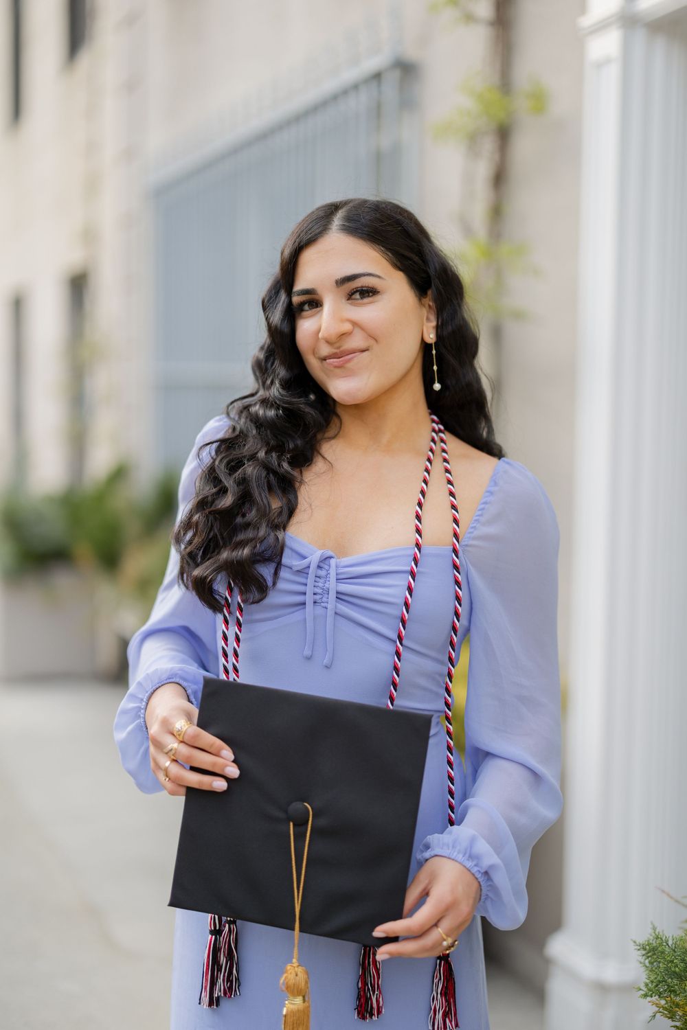 Graduate wearing periwinkle dress holds black graduation cap with gold tassel against light building exterior.