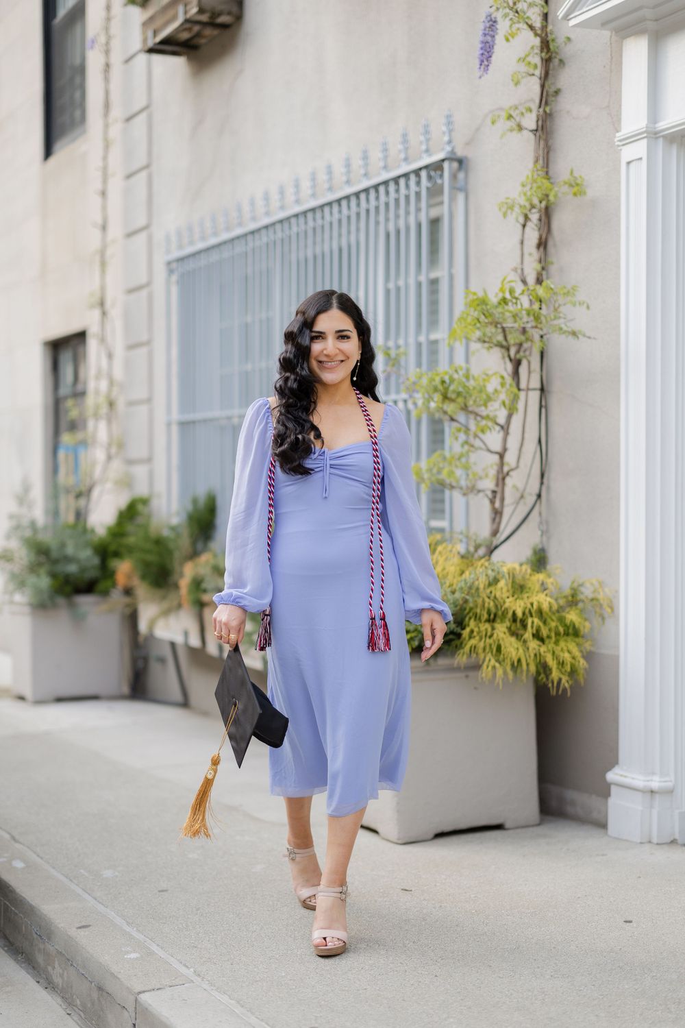 Graduate in flowing blue dress walks along sidewalk holding graduation cap with decorative tassels.