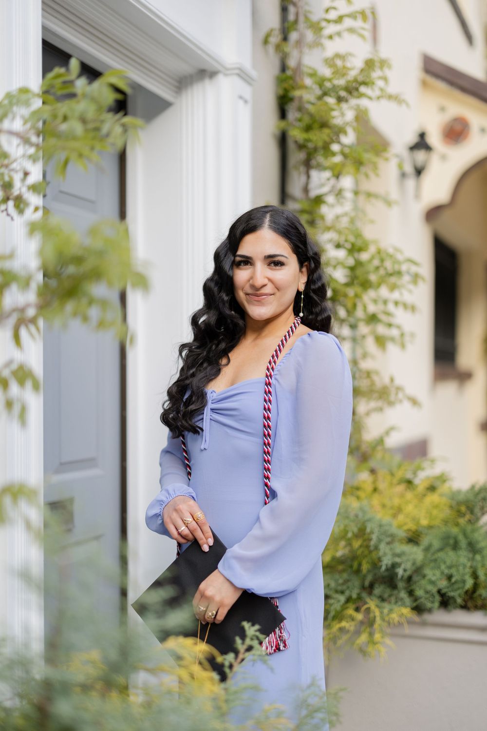Graduate in light blue dress poses with graduation cap against white building with greenery.