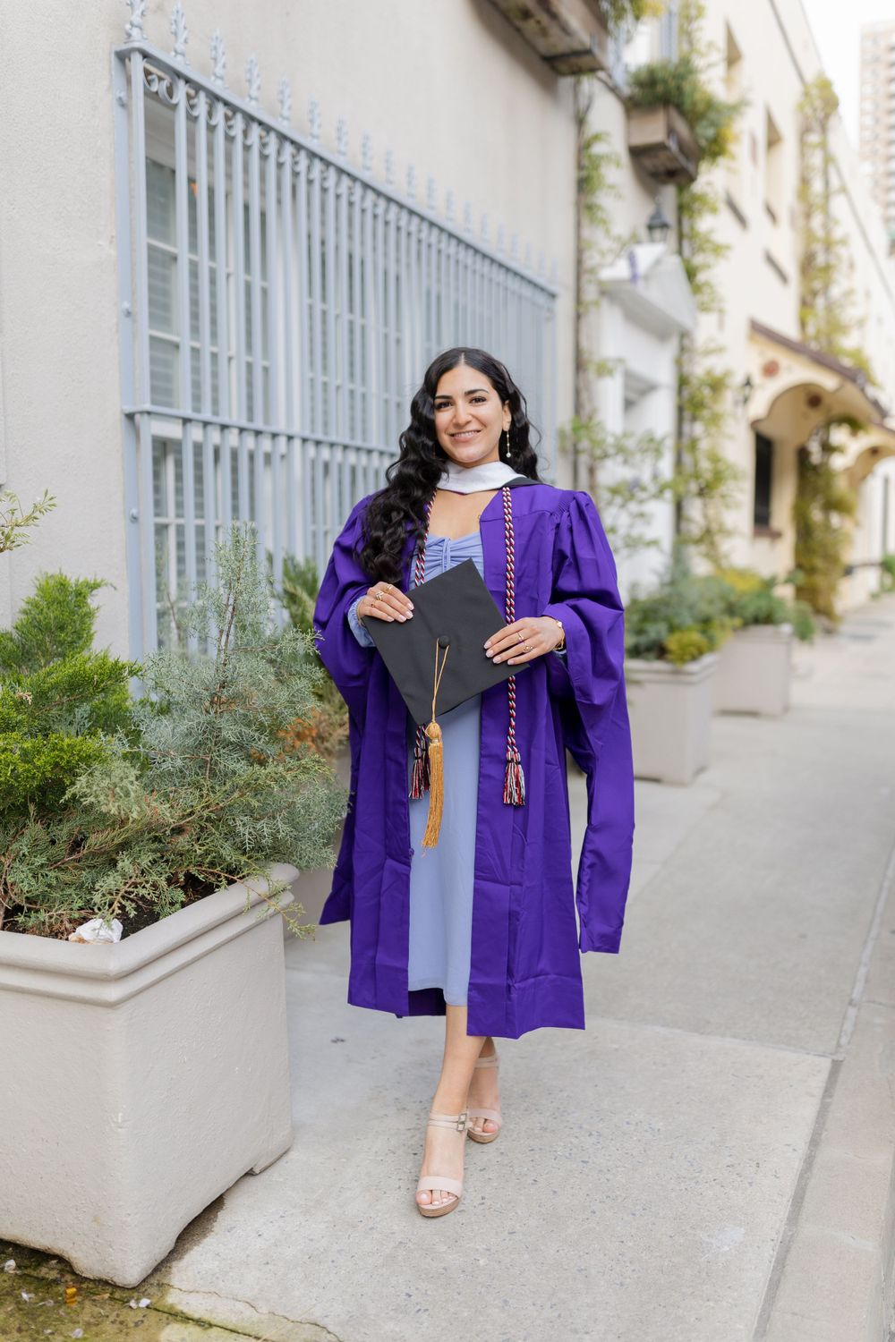 Graduate in purple graduation regalia and light blue dress poses with cap on city sidewalk.