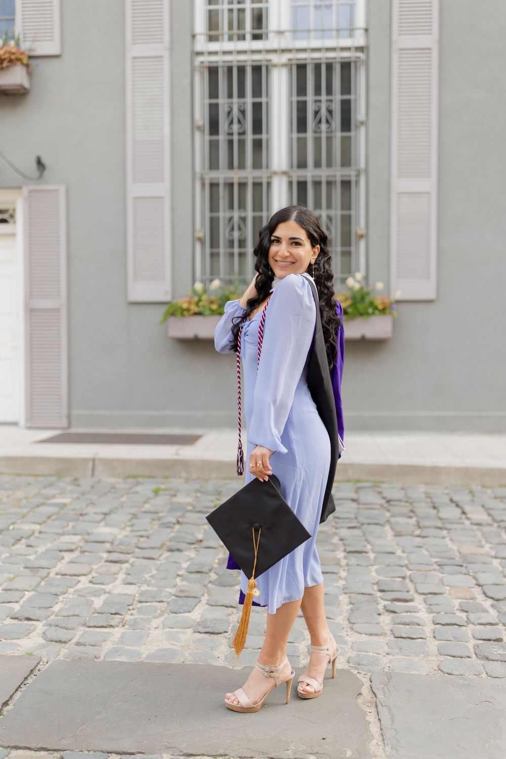 Graduate in periwinkle dress and purple gown poses with cap on cobblestone street.