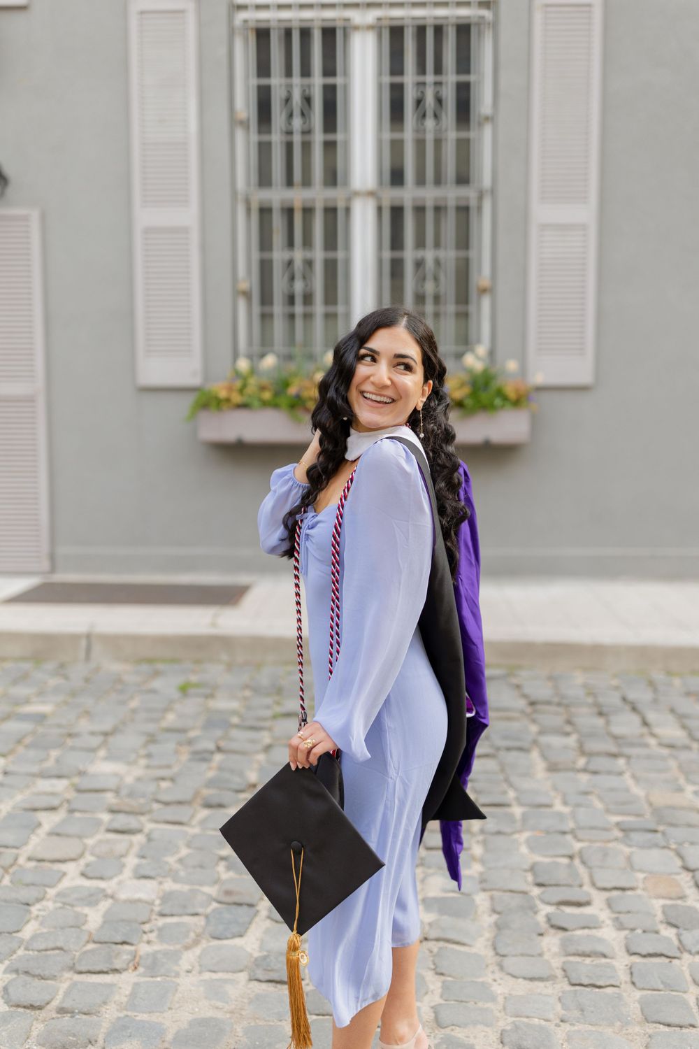 Graduate poses with graduation cap and gown on historic cobblestone street.