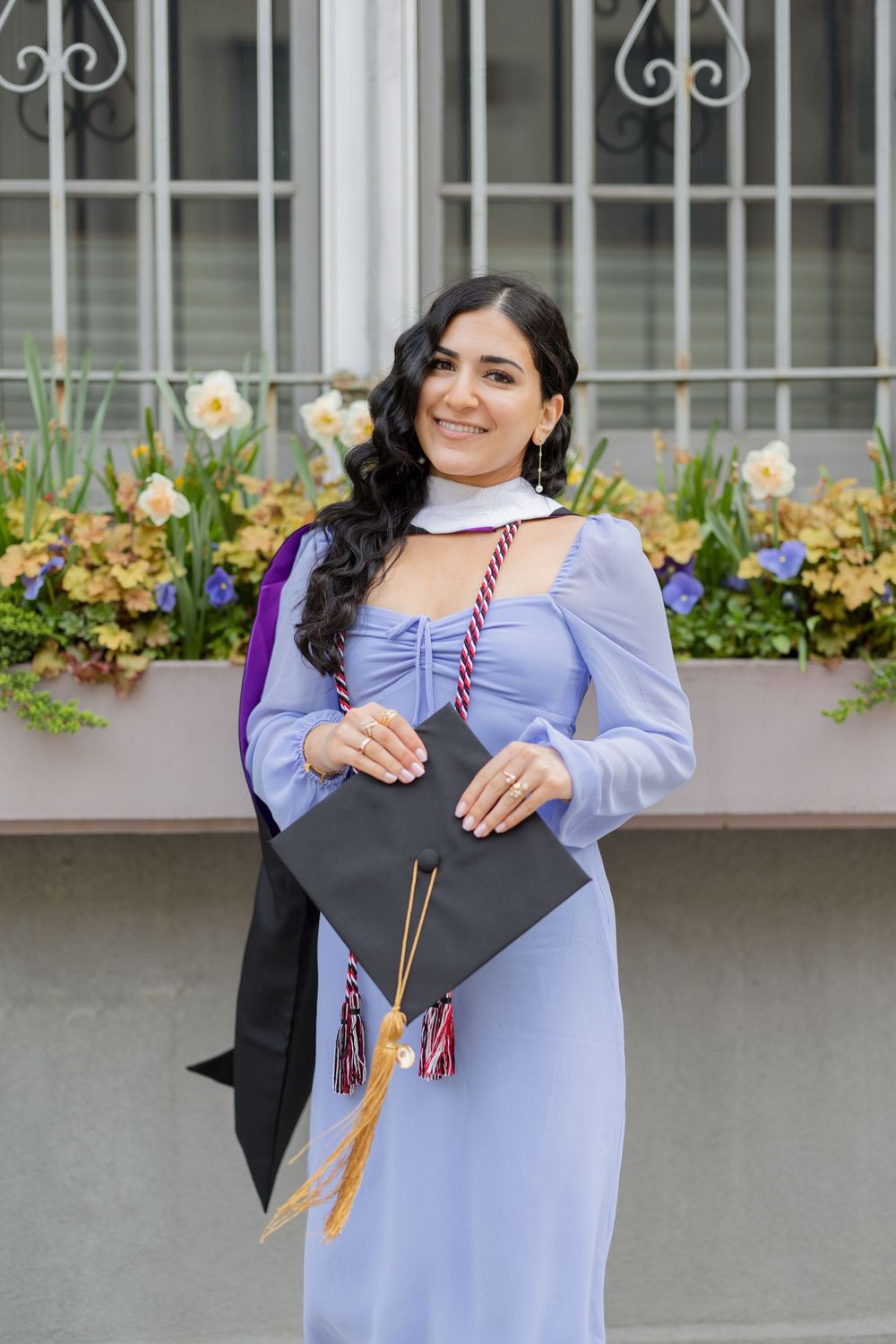 Graduate in light blue dress holds graduation cap in front of window box with spring flowers.