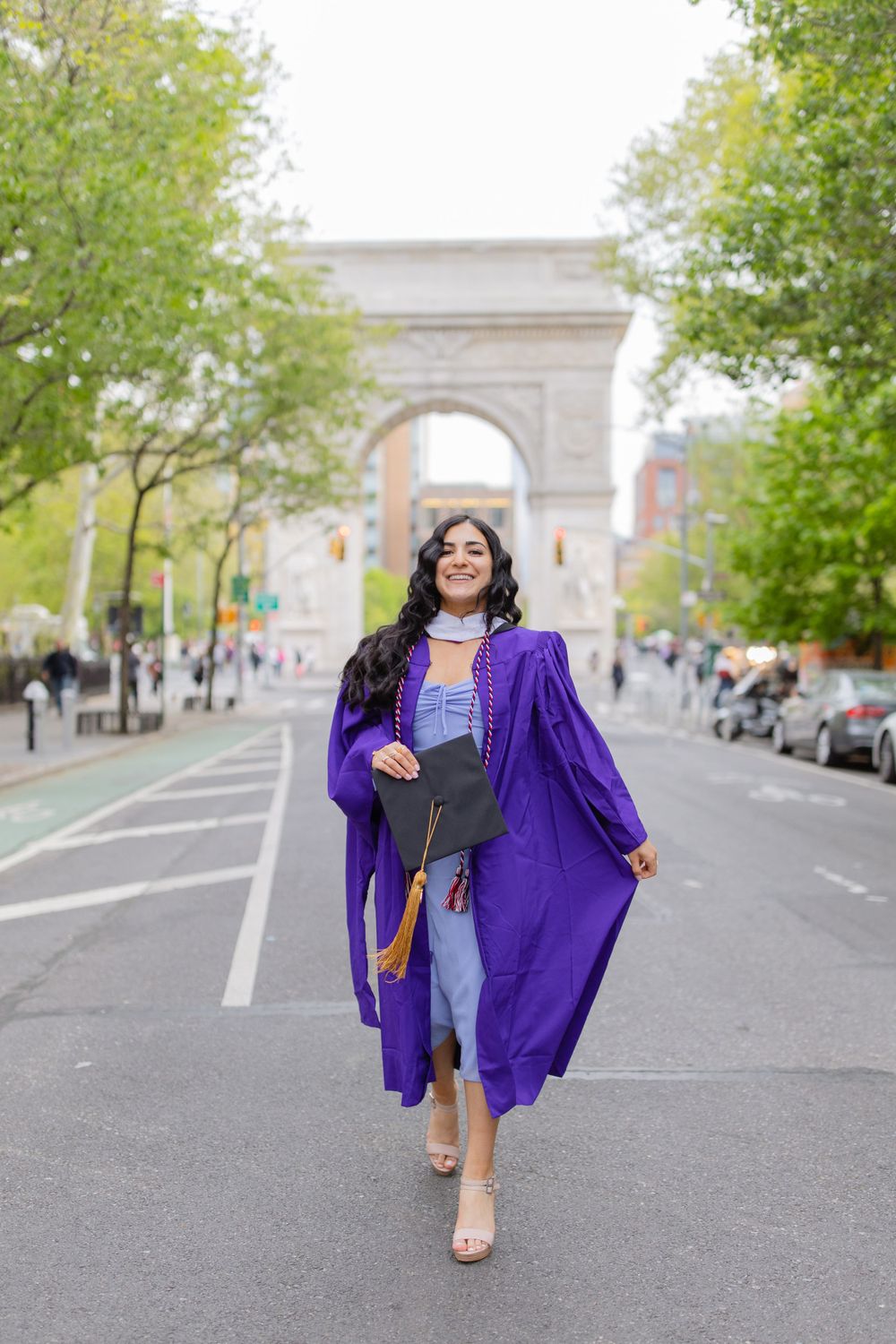 Graduate in purple graduation gown walks joyfully near Washington Square Arch.