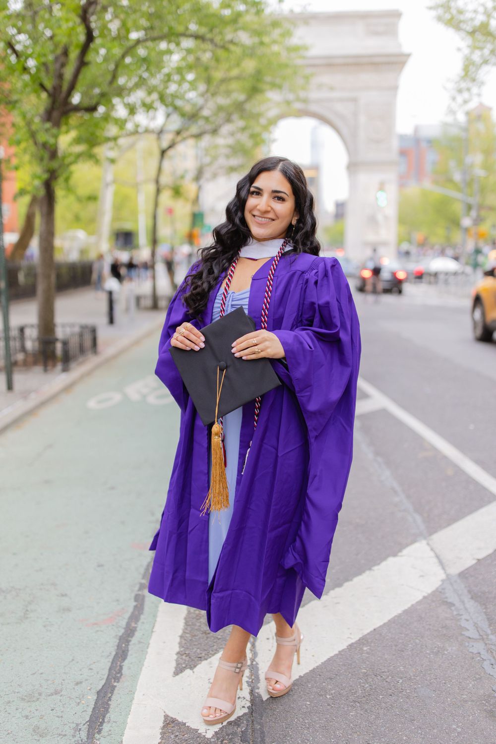 Graduate poses in purple graduation regalia with iconic Washington Square Arch in background.