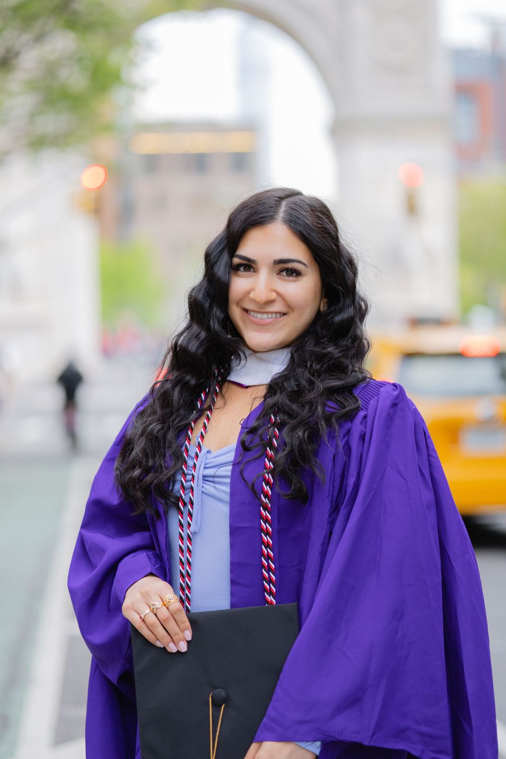 Graduate in purple academic regalia and light blue dress poses in front of Washington Square Arch.