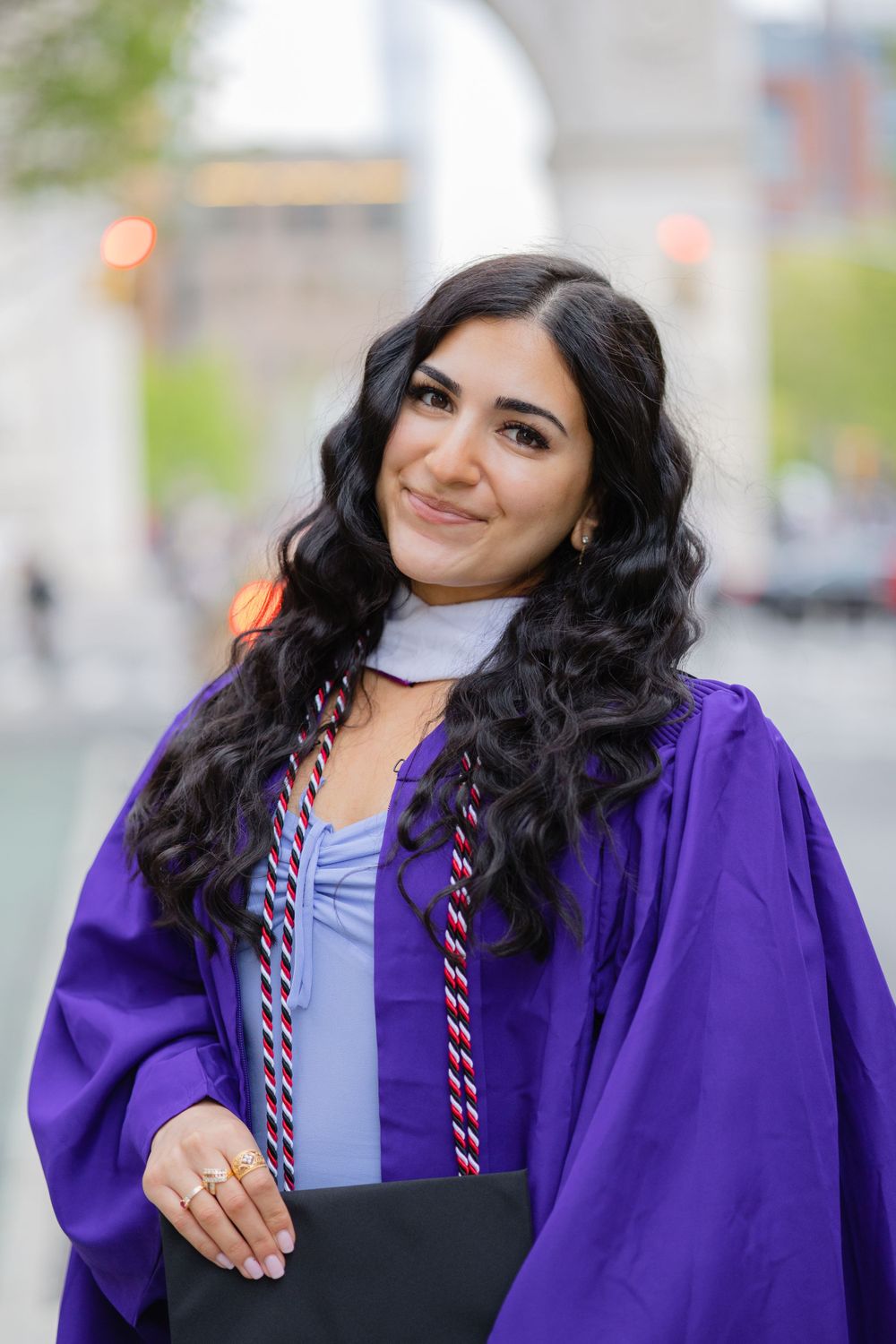 Graduate with wavy dark hair smiles while wearing purple graduation gown and honor cords.