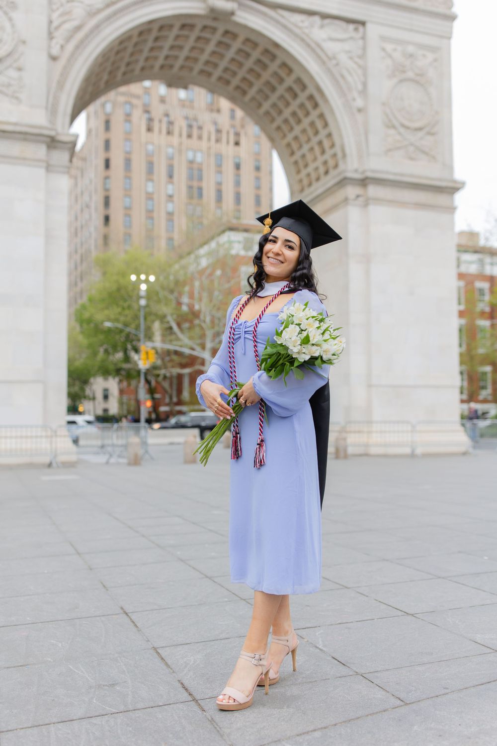 Graduate holds white flower bouquet while posing in academic regalia at Washington Square Park.