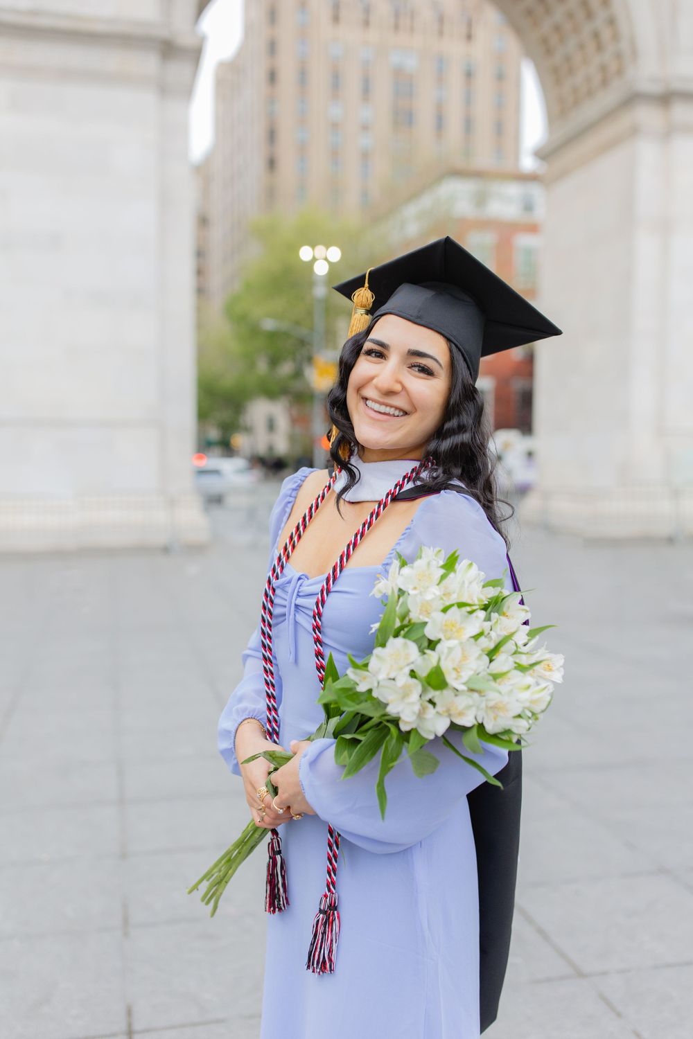Graduate poses with white flowers and graduation cap against iconic arch background.