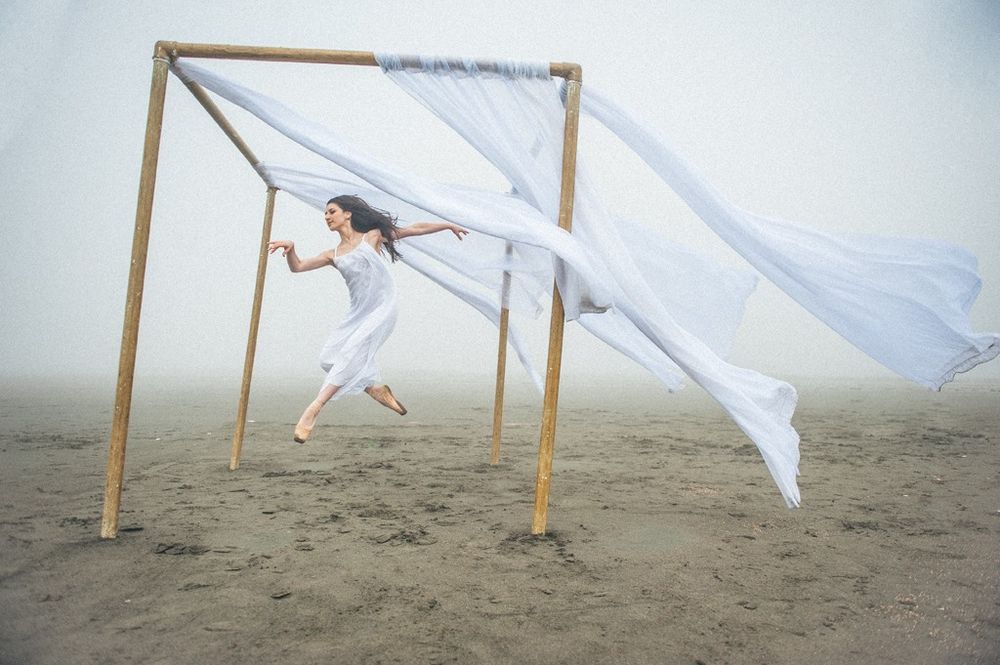 A figure in white dress dances beneath wooden beams on misty beach as flowing fabric billows in the wind.