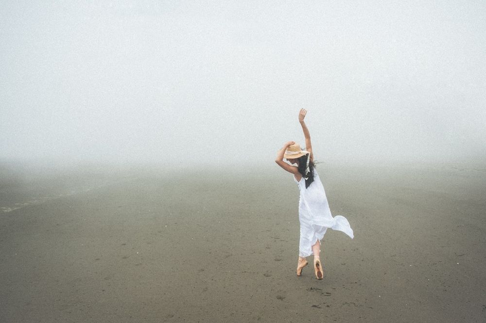 A figure in a flowing white dress leaps gracefully on a misty beach with ethereal fog in the background.