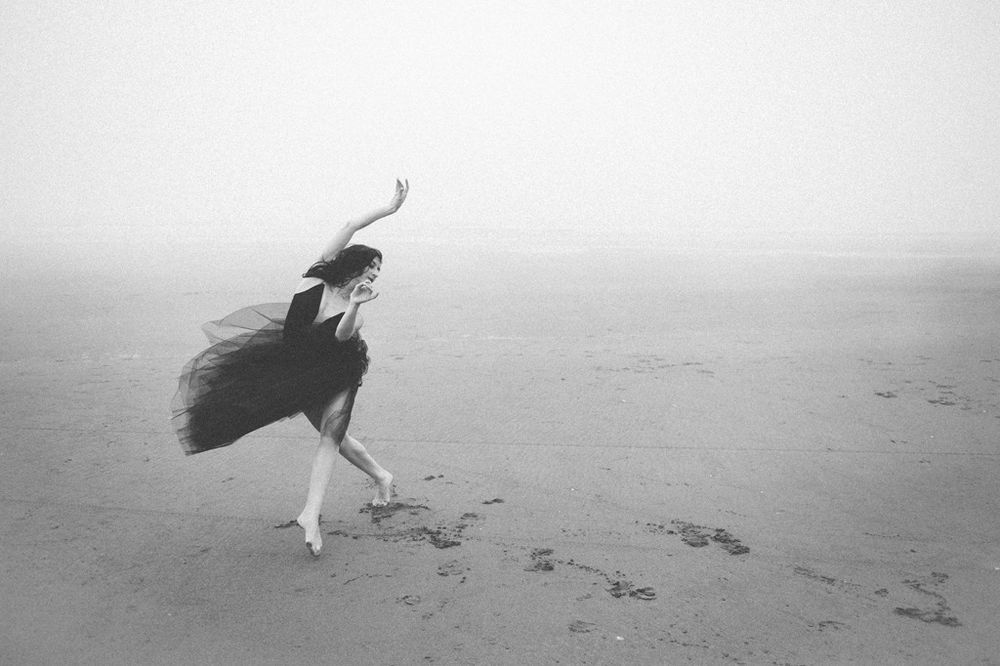 Black and white photo of a figure dancing on a foggy beach with dress and hair swept by wind.