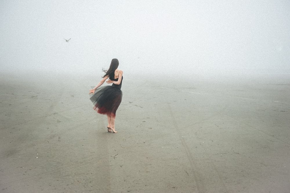 A person in a flowy black dress stands alone on a misty beach with their hair blowing in the wind.