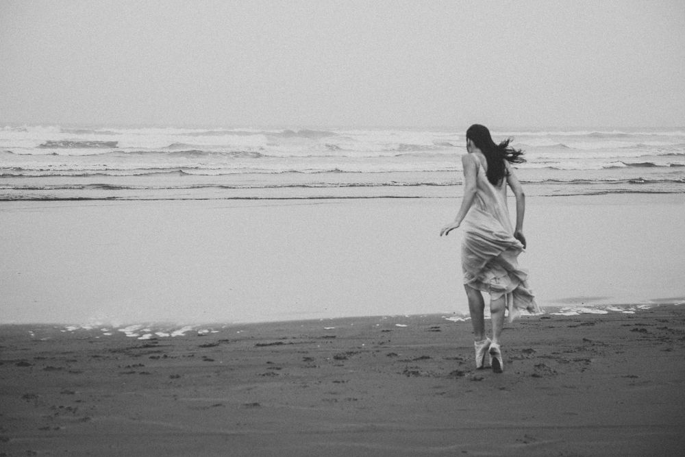 A person in flowing clothing walks alone on a misty beach shore in black and white.