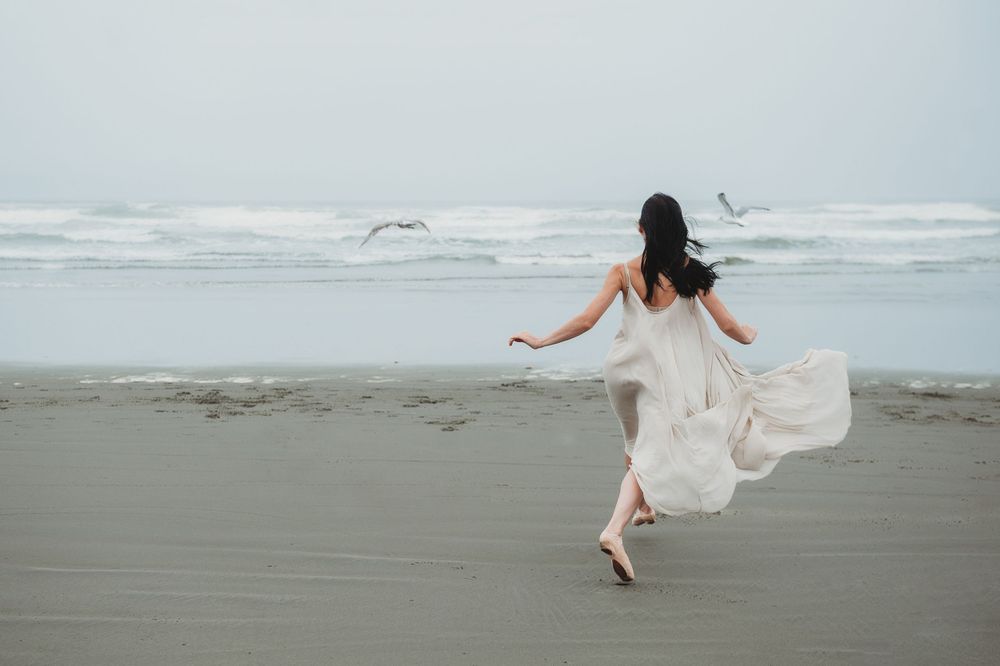 A flowing white dress catches the wind as a figure dances barefoot on a misty beach with seagulls flying overhead.