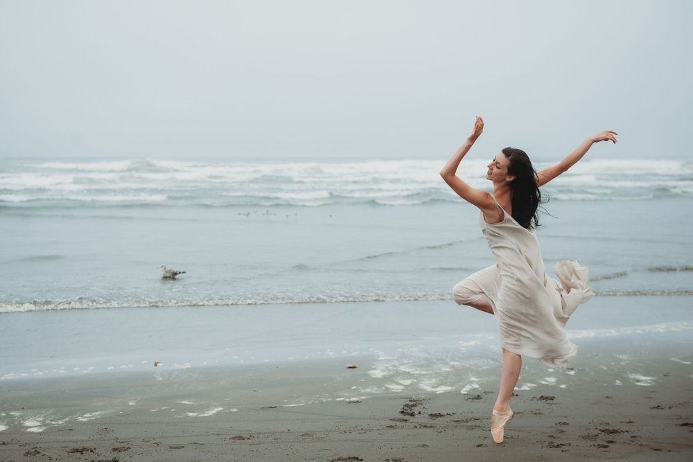 A dancer in a flowing white dress leaps gracefully on a misty beach with rolling waves in the background.