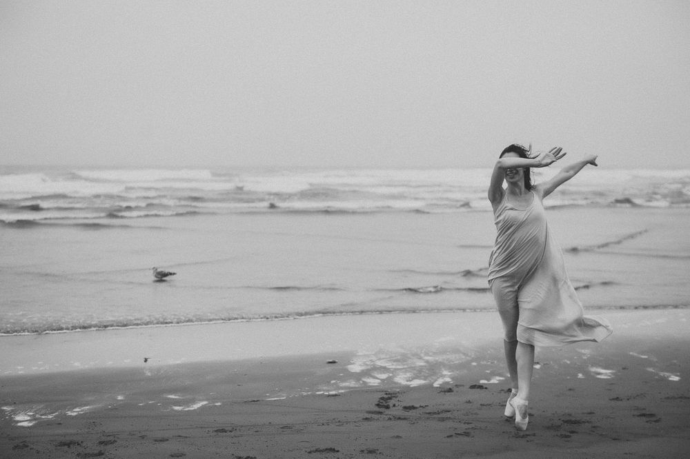 A graceful figure in a flowing white dress dances on a misty beach shoreline in black and white photography.