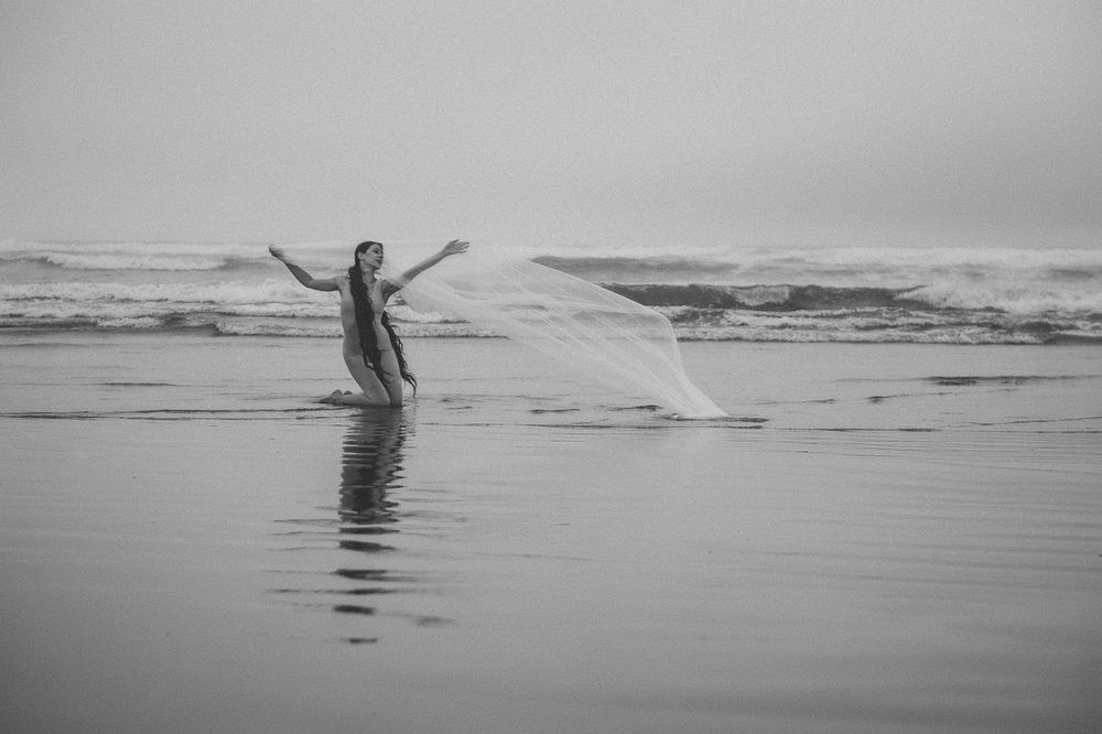 A figure in flowing fabric stands in shallow ocean water with arms outstretched in black and white.