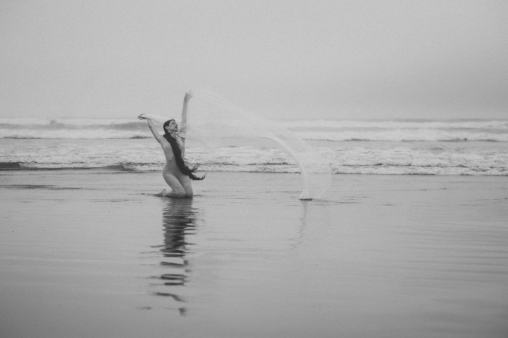 A figure silhouetted against ocean waves jumps joyfully on a misty beach in black and white photography.