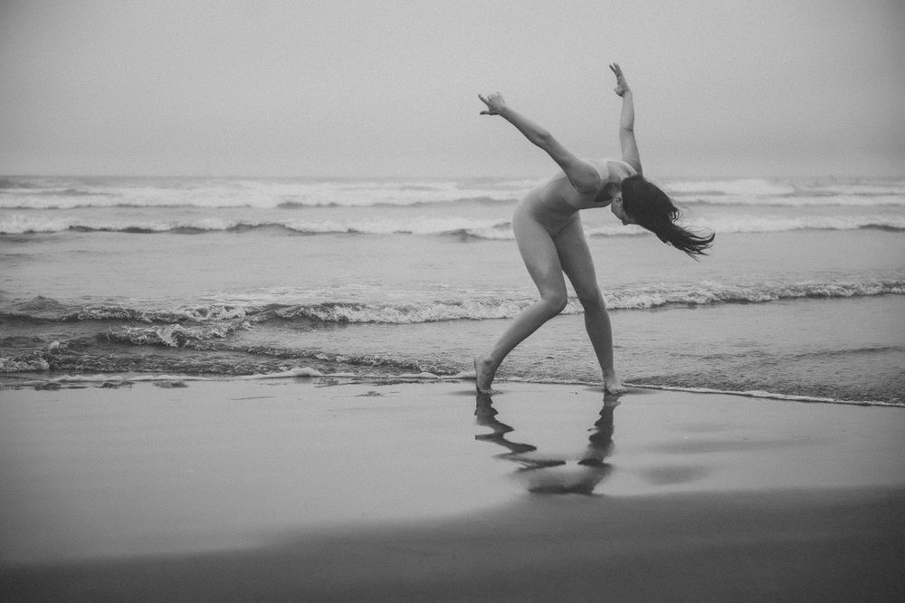 A graceful dancer performs a dynamic pose on the beach in black and white as waves crash in the background.