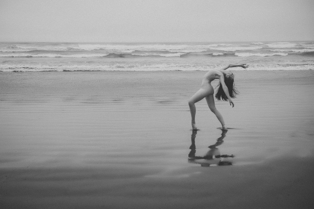 A graceful dancer performs on a misty beach with their reflection visible in the wet sand as waves roll in the background.
