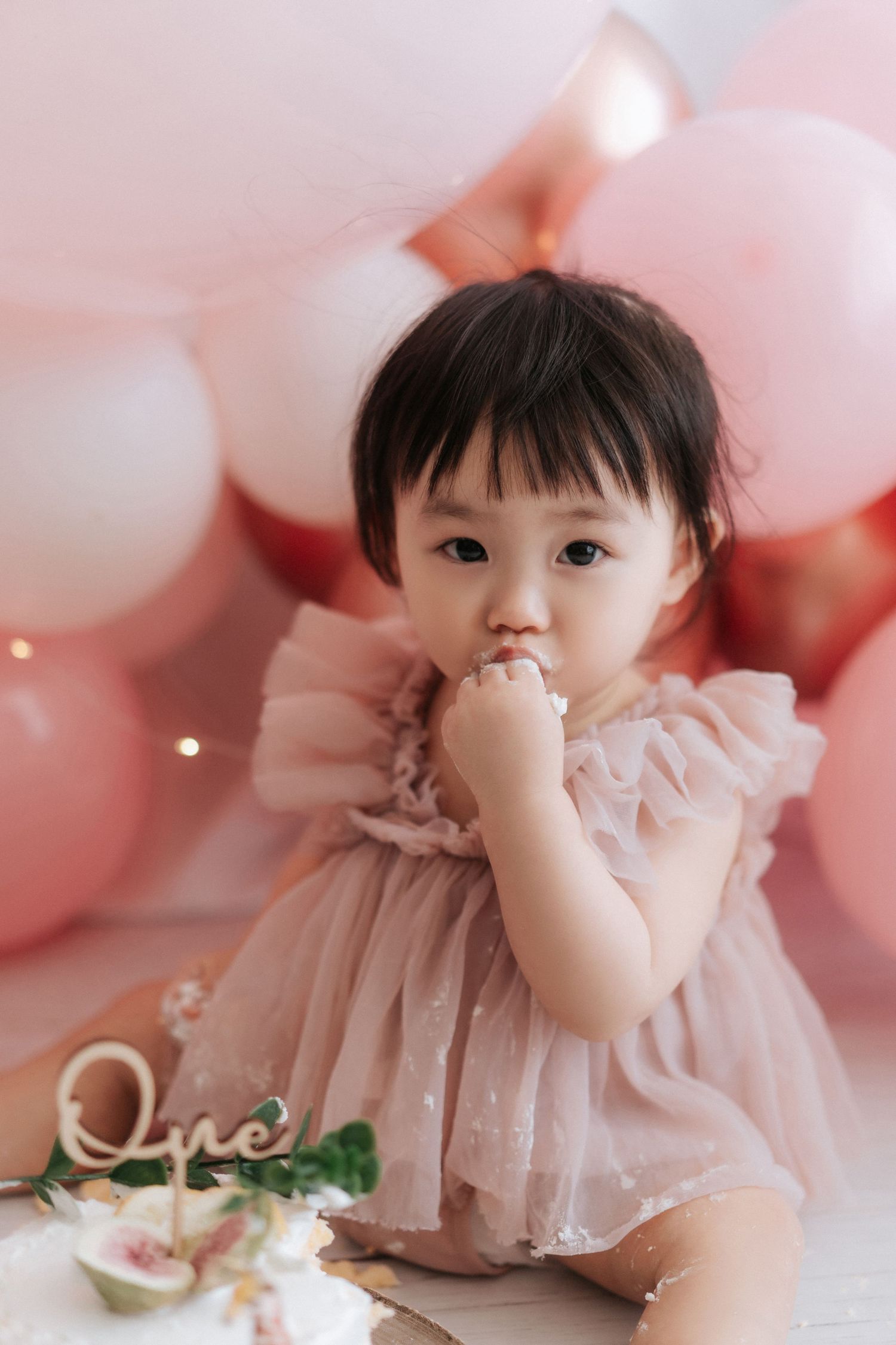 A baby in a pink ruffled dress poses sweetly on a light pink background with soft bokeh lighting effects.
