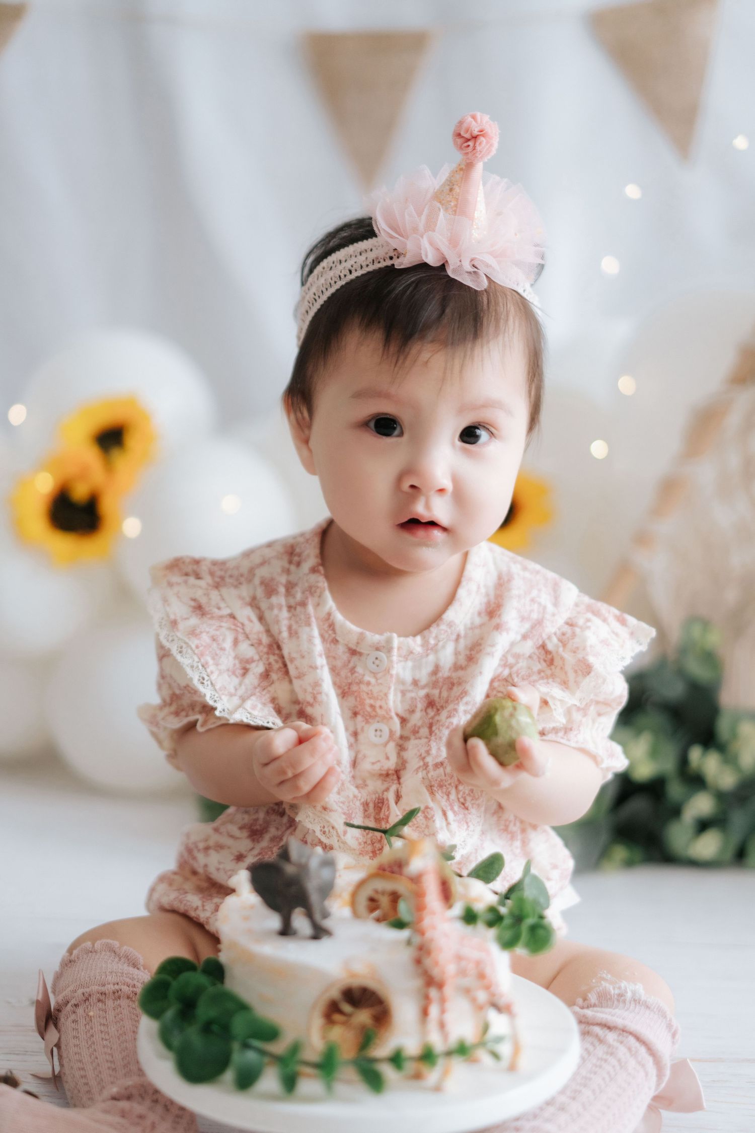 Baby in pink dress with crown headband sits with dinosaur-themed birthday cake decorated with toy figures and greenery.