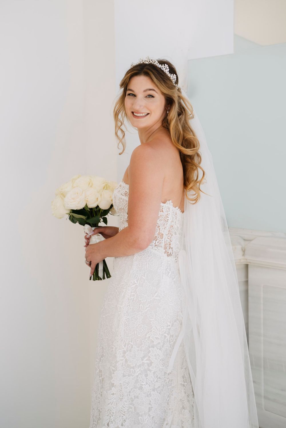 Bride in a stunning white lace wedding dress poses with a bouquet of white roses against a light background.