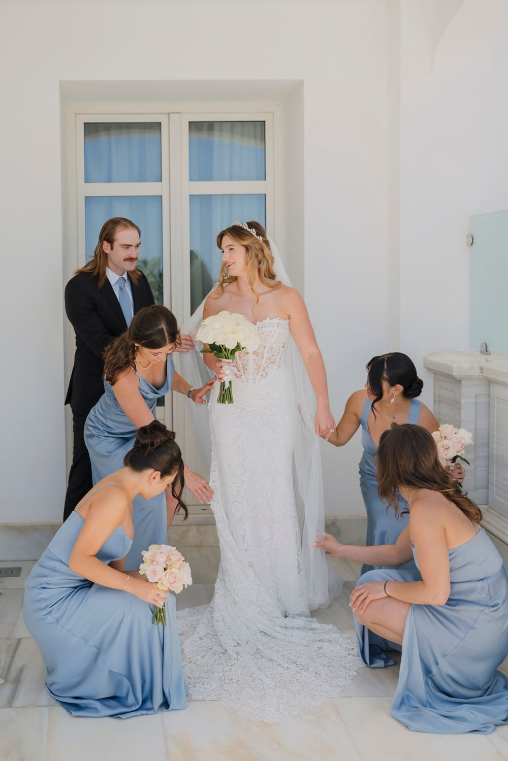 Bridesmaids in light blue dresses pose with the bride in a white gown against a neutral wall.