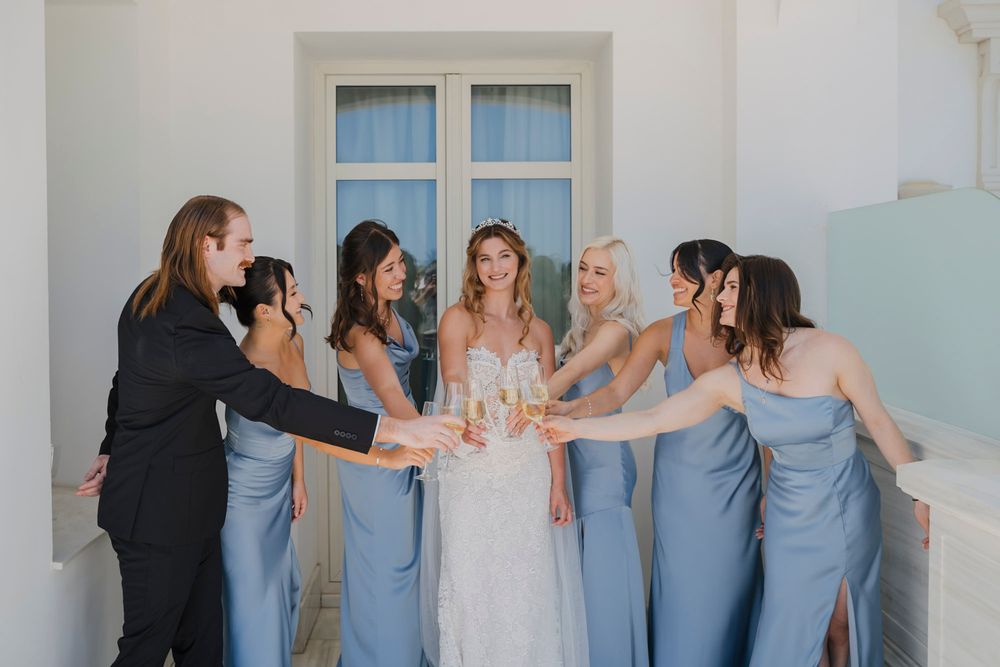 Wedding party in light blue dresses poses together against a white wall in a candid moment.